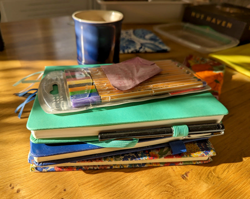 Pile of sketchbooks in a sunbeam, waiting for ideas! With thin markers and a cup of tea in background